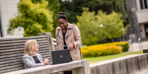 Two individuals outside talking and looking at a laptop, one standing one sitting